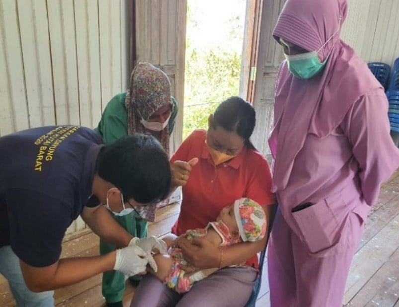 A baby getting immunization while being held by a woman. Three health workers stand around the woman.
